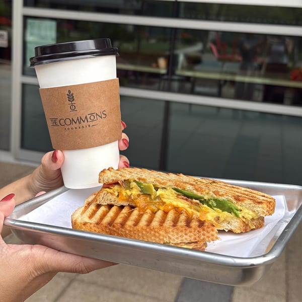 A hand holds a Commons Fooderie coffee cup and a panini with egg, cheese, and avocado.