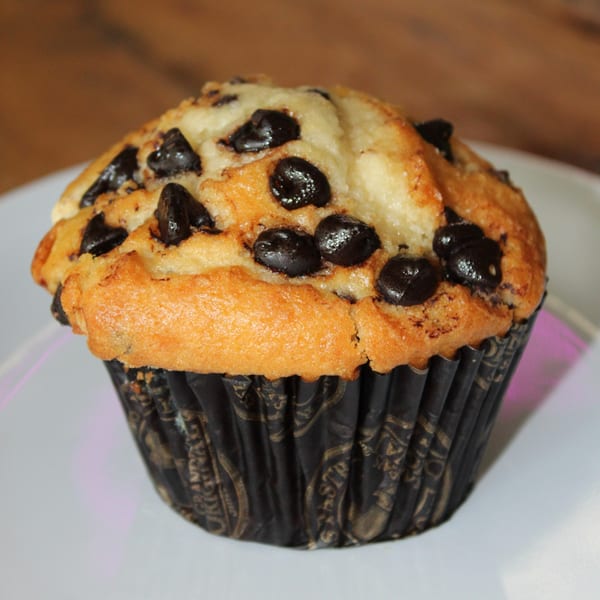 A golden-brown chocolate chip muffin in a dark wrapper sits on a white plate.