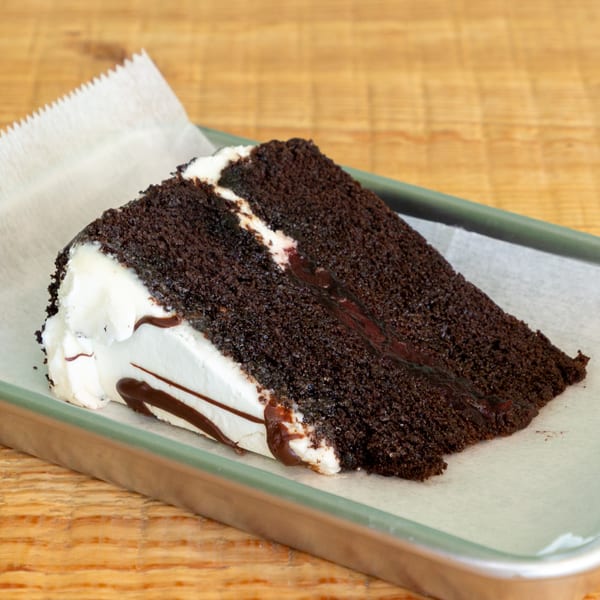 A slice of rich chocolate cake with white frosting and chocolate drizzle on a metal tray.
