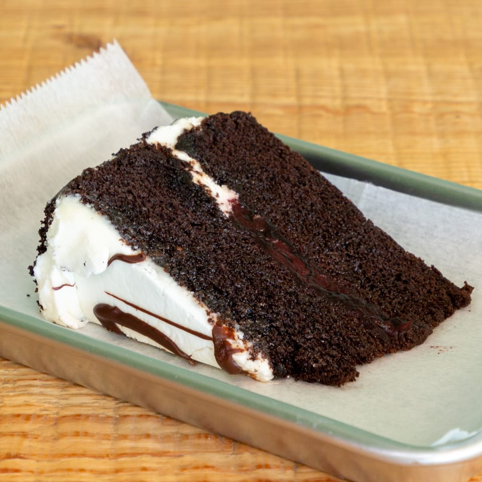 A slice of rich chocolate cake with white frosting and chocolate drizzle on a metal tray.