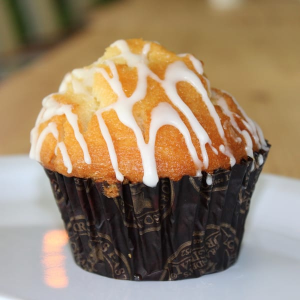 A golden muffin with white icing drizzle, in a dark patterned wrapper, sits on a white plate.