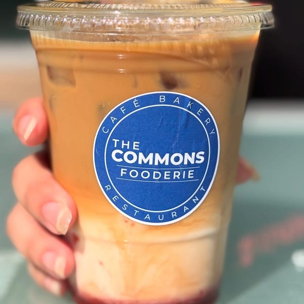 Hand holding a clear plastic cup of iced coffee with the blue "The Commons Fooderie" logo.