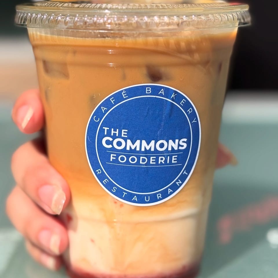 Hand holding a clear plastic cup of iced coffee with the blue "The Commons Fooderie" logo.
